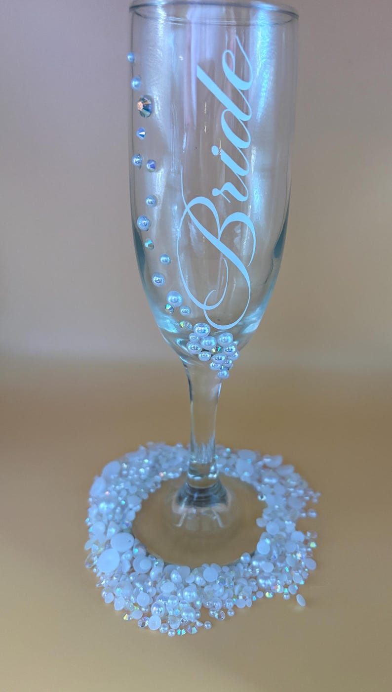 May include: A clear champagne flute with the word "Bride" in elegant script. The glass is adorned with small, sparkling embellishments and a base ringed with white beads, creating a celebratory and decorative design.