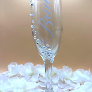 Puede incluir: Una copa de champ&aacute;n transparente con la palabra &laquo;Bride&raquo; en escritura azul claro. El vaso est&aacute; adornado con peque&ntilde;as cuentas iridiscentes y adornos redondos y transparentes. La copa descansa sobre un lecho de p&eacute;talos de flores blancas.