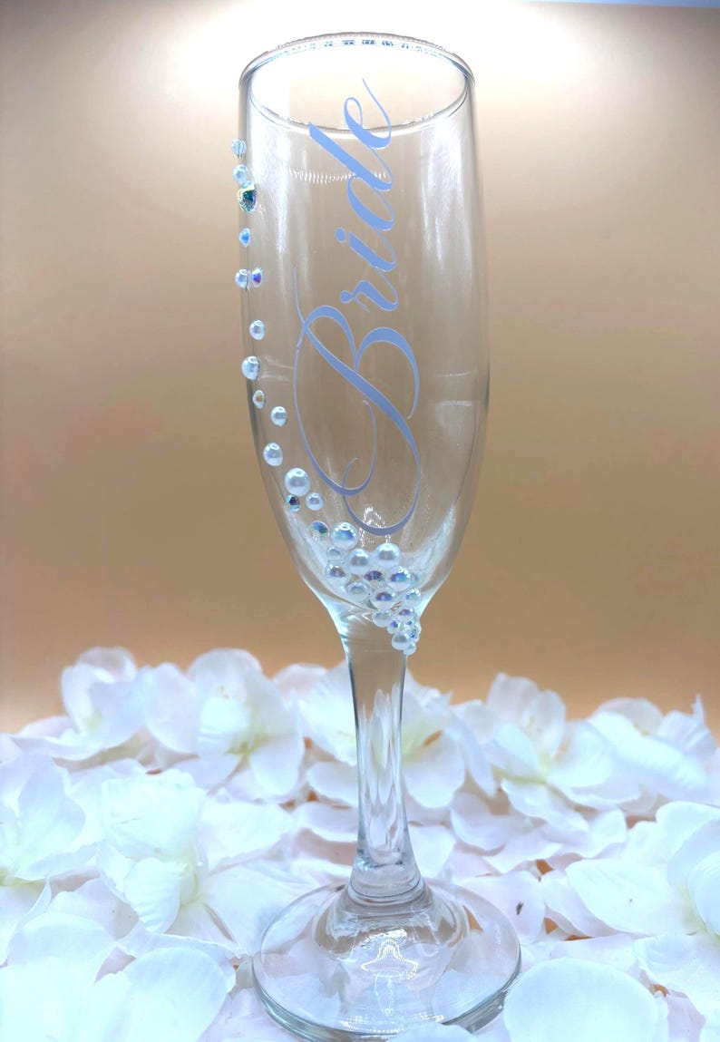 Puede incluir: Una copa de champ&aacute;n transparente con la palabra &laquo;Bride&raquo; en escritura azul claro. El vaso est&aacute; adornado con peque&ntilde;as cuentas iridiscentes y adornos redondos y transparentes. La copa descansa sobre un lecho de p&eacute;talos de flores blancas.
