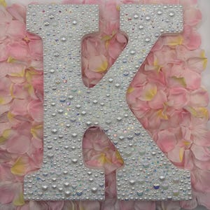 May include: A large, white letter "K" is covered in iridescent beads and pearls. The letter is set against a backdrop of pink and yellow flower petals, creating a soft, feminine aesthetic. This decorative piece is ideal for personalized decor.