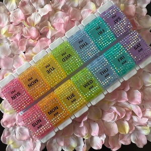 Puede incluir: Un organizador de pastillas para siete d&iacute;as con compartimentos para cada d&iacute;a de la semana, etiquetados con los d&iacute;as de la semana y AM o PM. Los compartimentos est&aacute;n decorados con brillantes de colores.