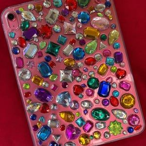 Könnte beinhalten: Eine rosa Tablet-Hülle, verziert mit einer Ansammlung farbenfroher, funkelnder Edelsteine. Die Edelsteine haben verschiedene Formen und Größen, darunter Quadrate, Ovale und Herzen, und sind in den Farbtönen Rot, Blau, Grün, Gelb und Lila gehalten.