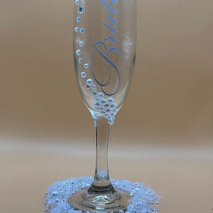 Puede incluir: Una copa de champ&aacute;n transparente con la palabra "Bride" en escritura azul claro. El vaso est&aacute; decorado con pedrer&iacute;a iridiscente y un grupo de pedrer&iacute;a en la base. Un anillo de pedrer&iacute;a blanca rodea la base del vaso.