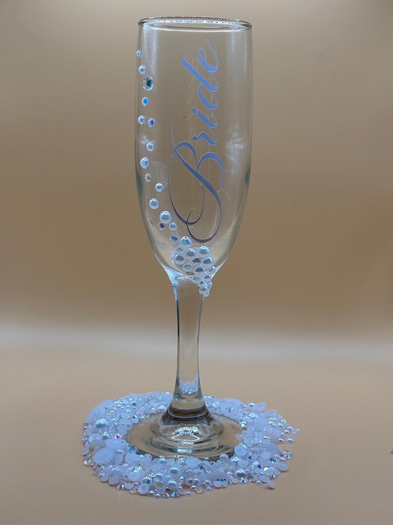 Puede incluir: Una copa de champ&aacute;n transparente con la palabra "Bride" en escritura azul claro. El vaso est&aacute; decorado con pedrer&iacute;a iridiscente y un grupo de pedrer&iacute;a en la base. Un anillo de pedrer&iacute;a blanca rodea la base del vaso.