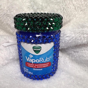 Könnte beinhalten: Ein verziertes Glas Vicks VapoRub Salbe. Das blaue Glas ist mit blauen Strasssteinen verziert, und der Deckel hat einen grünen Strasssteinrand. Das Etikett lautet "Vicks VapoRub COUGH SUPPRESSANT TOPICAL ANALGESIC Ointment" und "NET WT 100 g".