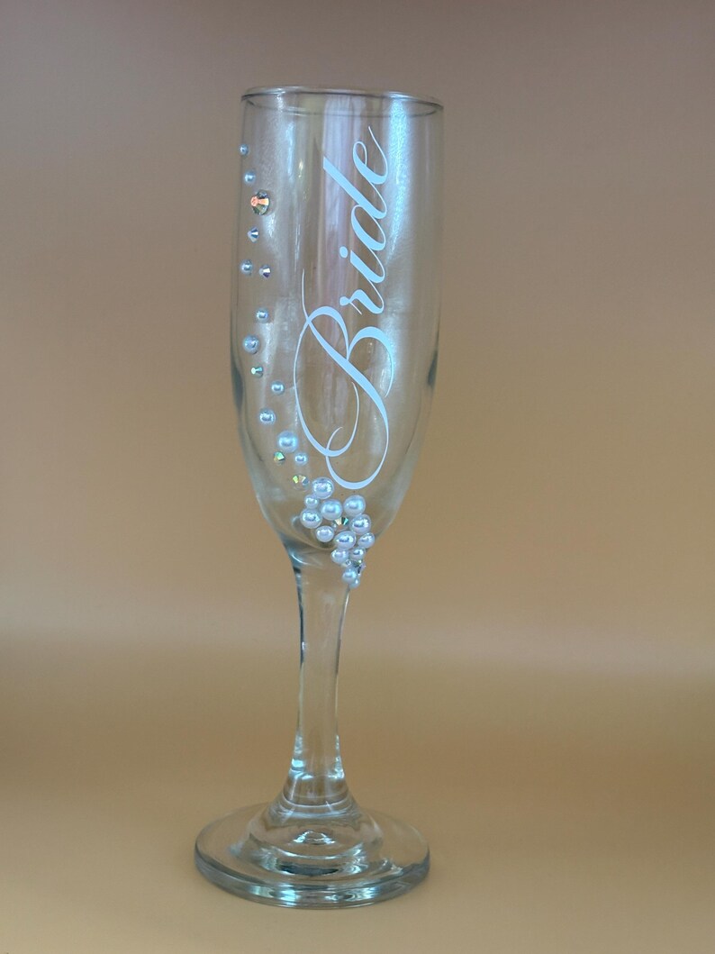 May include: A clear champagne flute with the word "Bride" in elegant script. The glass is adorned with small, pearl-like embellishments. The stem and base are also clear, creating a delicate and celebratory aesthetic.