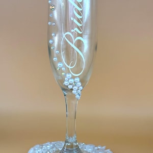 Puede incluir: Una copa de champ&aacute;n transparente con la palabra "Bride" en escritura elegante. El vaso est&aacute; adornado con peque&ntilde;os pedrer&iacute;a y perlas de imitaci&oacute;n. La base del vaso est&aacute; rodeada por un lecho de perlas blancas y gemas iridiscentes.