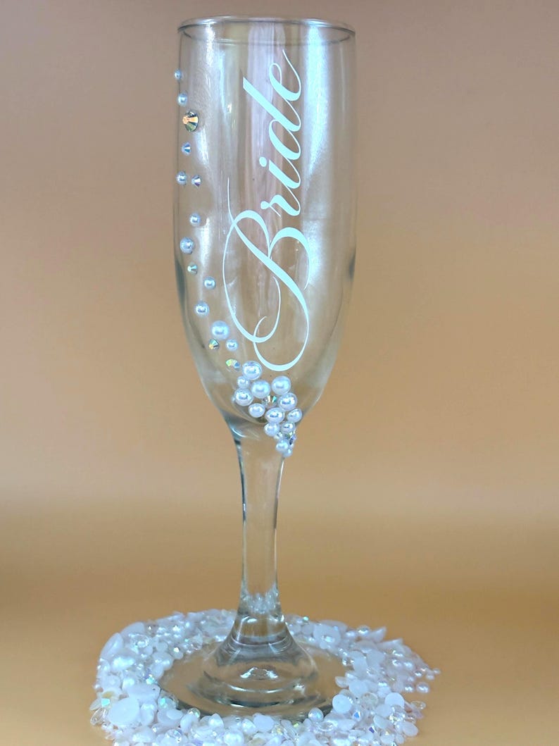 Puede incluir: Una copa de champ&aacute;n transparente con la palabra "Bride" en escritura elegante. El vaso est&aacute; adornado con peque&ntilde;os pedrer&iacute;a y perlas de imitaci&oacute;n. La base del vaso est&aacute; rodeada por un lecho de perlas blancas y gemas iridiscentes.