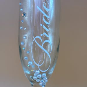 Puede incluir: Una copa de champ&aacute;n transparente con la palabra "Bride" en escritura azul claro. El vaso est&aacute; decorado con peque&ntilde;as cuentas iridiscentes de color azul claro y un solo cristal m&aacute;s grande. Un art&iacute;culo con tem&aacute;tica de boda.