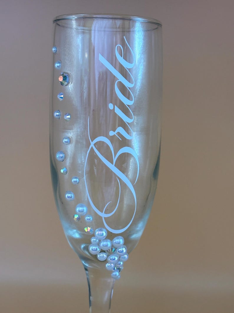 Puede incluir: Una copa de champ&aacute;n transparente con la palabra "Bride" en escritura azul claro. El vaso est&aacute; decorado con peque&ntilde;as cuentas iridiscentes de color azul claro y un solo cristal m&aacute;s grande. Un art&iacute;culo con tem&aacute;tica de boda.