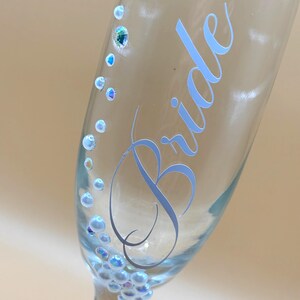 Puede incluir: Una copa de champ&aacute;n transparente con la palabra "Bride" en escritura azul claro. El vaso est&aacute; adornado con adornos iridiscentes en forma de perlas y peque&ntilde;os cristales. El dise&ntilde;o es elegante y adecuado para una celebraci&oacute;n de boda.