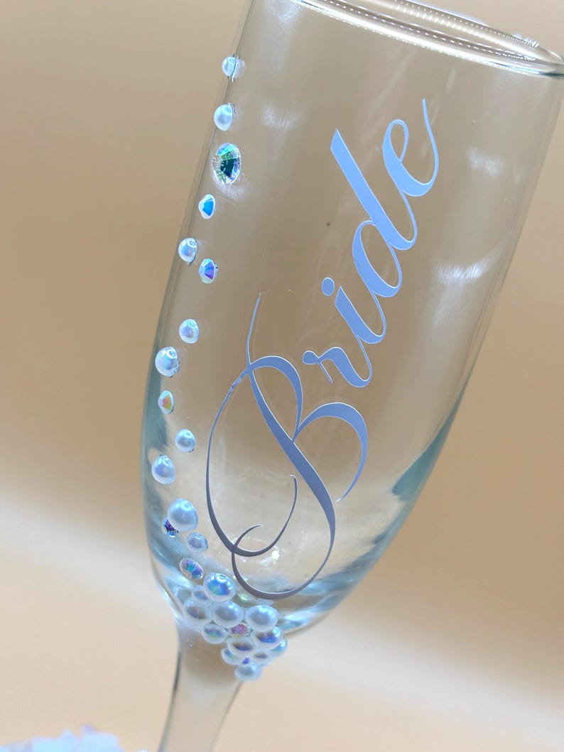 Puede incluir: Una copa de champ&aacute;n transparente con la palabra "Bride" en escritura azul claro. El vaso est&aacute; adornado con adornos iridiscentes en forma de perlas y peque&ntilde;os cristales. El dise&ntilde;o es elegante y adecuado para una celebraci&oacute;n de boda.