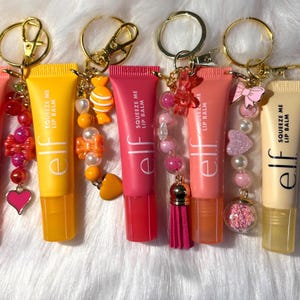 May include: A selection of 'elf' brand lip balm tubes, each attached to a keychain. The tubes are in various shades like pink and yellow, and are decorated with charms. The text "Squeeze Me Lip Balm" is printed on each tube, adding a touch of detail.