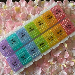 Puede incluir: Un organizador de pastillas de pl&aacute;stico transparente con un arco&iacute;ris de colores y brillantes. Cada compartimento est&aacute; etiquetado con el d&iacute;a de la semana y AM o PM. El organizador est&aacute; sentado sobre un lecho de p&eacute;talos de flores rosas.