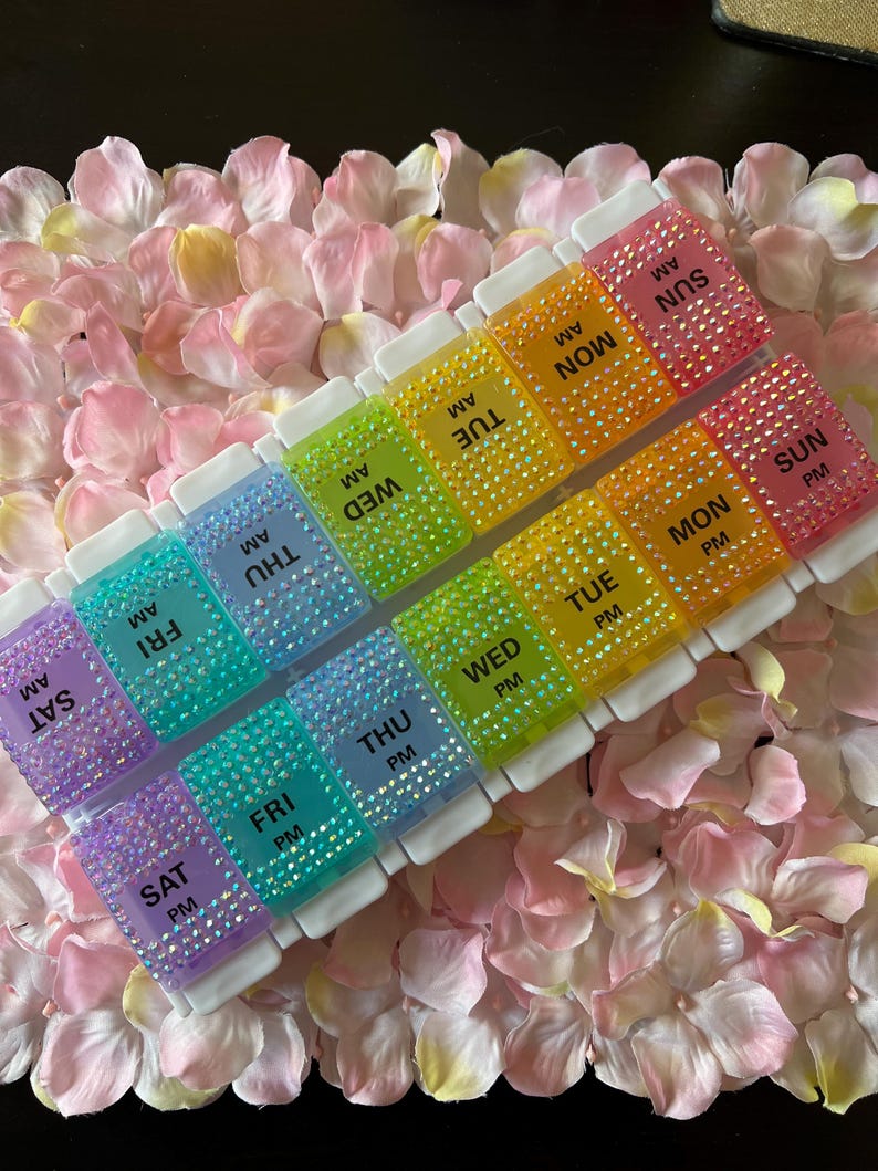 Puede incluir: Un organizador de pastillas de pl&aacute;stico transparente con un arco&iacute;ris de colores y brillantes. Cada compartimento est&aacute; etiquetado con el d&iacute;a de la semana y AM o PM. El organizador est&aacute; sentado sobre un lecho de p&eacute;talos de flores rosas.