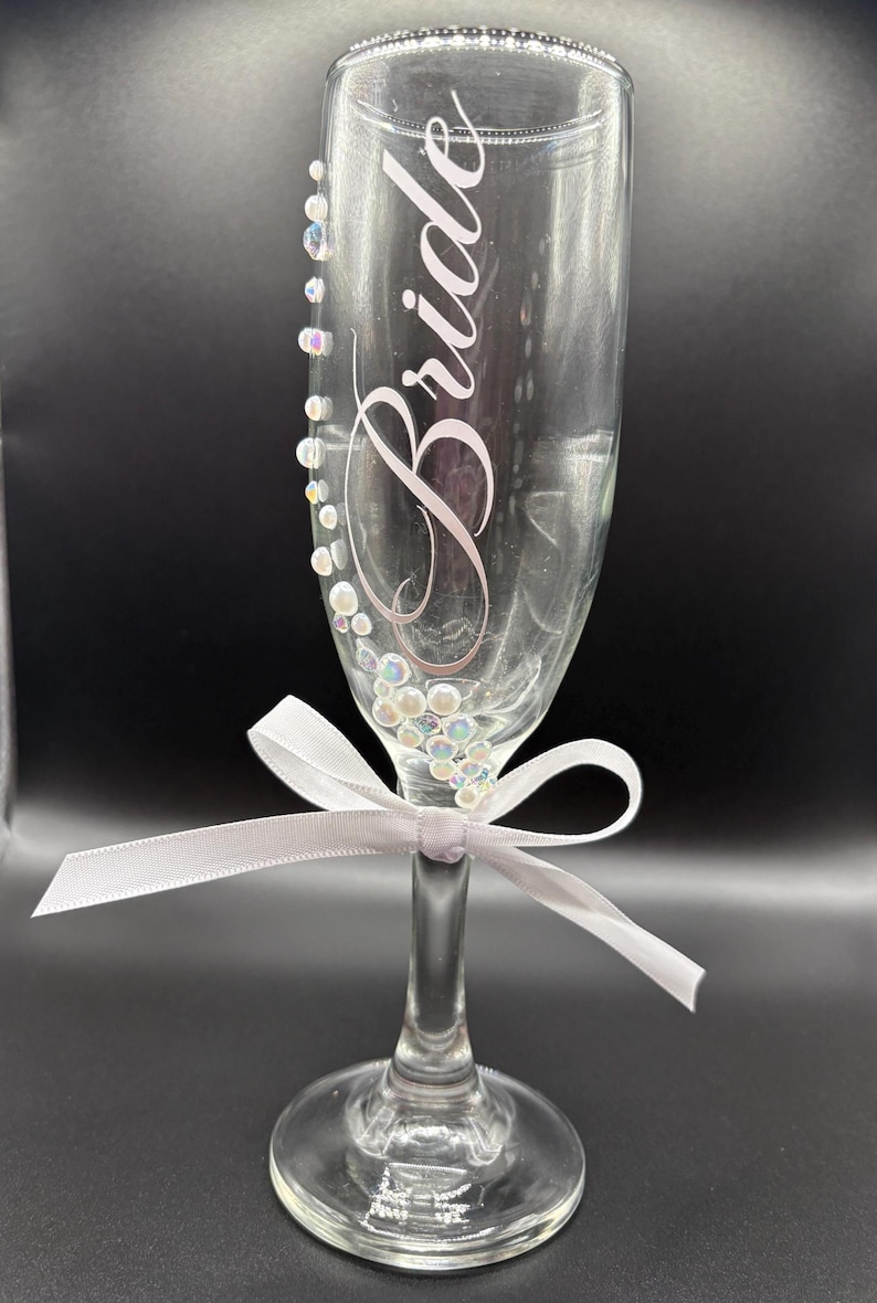 Puede incluir: Una flauta de champ&aacute;n transparente con la palabra "Bride" en cursiva. El vaso est&aacute; adornado con pedrer&iacute;a iridiscente y una cinta blanca atada alrededor del tallo. El vaso est&aacute; dise&ntilde;ado para una boda.