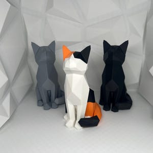 May include: Three origami cat figurines, one black, one gray, and one calico with orange, black, and white. The cats are sitting on a white background.