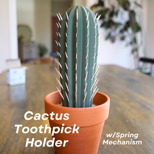 May include: A green cactus-shaped toothpick holder with a brown terracotta pot. The holder has a spring mechanism and the text "Cactus Toothpick Holder w/Spring Mechanism" is printed on the table below the holder.
