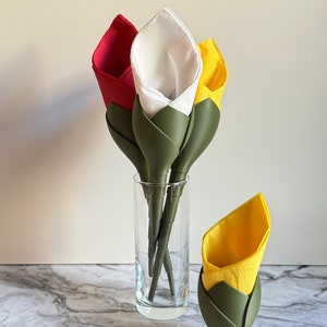 May include: A clear glass vase holds three decorative tulip-shaped napkins in red, white, and yellow. A single yellow tulip napkin sits to the side. The napkins are folded to resemble flowers, with green stems. The background is a marble-patterned surface.