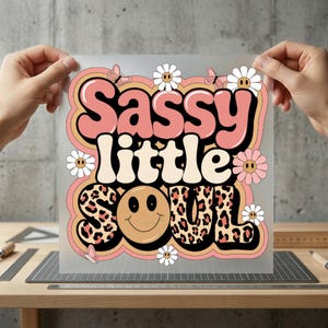 May include: A transparent sheet with the words "Sassy little soul" in a retro design. The text is in pink, cream, and leopard print, with a smiley face and flower accents. The sheet is held by two hands.