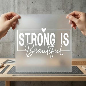 May include: A frosted, transparent sheet with the words "STRONG IS Beautiful" in white script and block letters, framed by a white rectangle and a small heart above the word "IS". The sheet is held up by two hands.