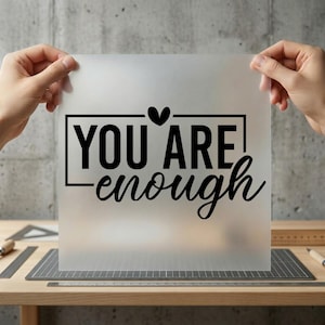 May include: A frosted sheet with the words "YOU ARE enough" in black script and block letters, with a heart symbol above the word "ARE". The sheet is held up by two hands against a neutral background.