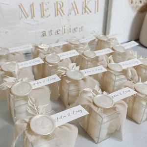 Puede incluir: Una colección de pequeñas cajas cuadradas transparentes llenas de caramelos blancos. Cada caja está envuelta en una tela beige con un sello de cera blanco y una pequeña rama de vegetación seca. Las cajas están etiquetadas con el texto "Jelin & Eray".