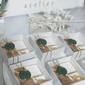 May include: White gift boxes with gold ribbon and a green wax seal. Each box contains a small brown paper bag with gold lettering and a gold spoon. The boxes are arranged in a row on a white surface. The text on the boxes reads "Enhan & Cansin".