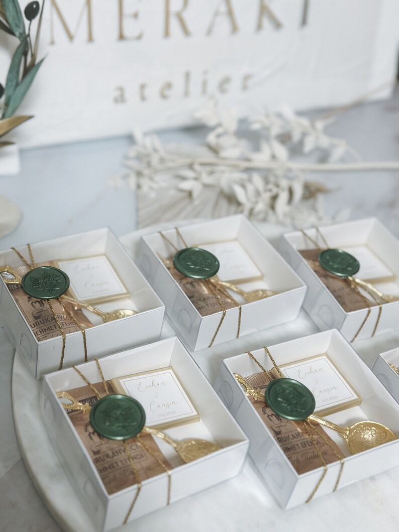 May include: White gift boxes with gold ribbon and a green wax seal. Each box contains a small brown paper bag with gold lettering and a gold spoon. The boxes are arranged in a row on a white surface. The text on the boxes reads "Enhan & Cansin".