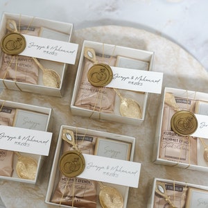 May include: A collection of small, white gift boxes with gold accents. Each box contains a small bag of coffee, a gold spoon, and a wax seal with a ring design. The boxes are labeled with the names "Surayya & Muhammed" and the date "11.10.2023".