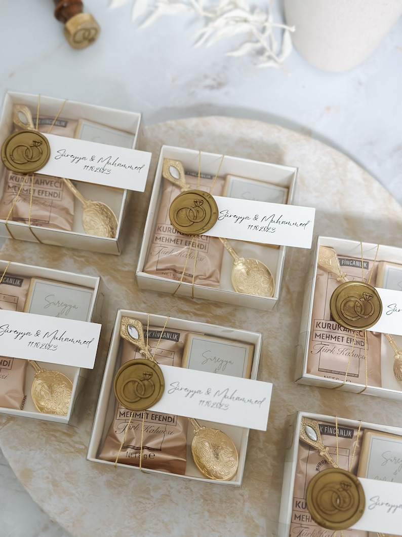 May include: A collection of small, white gift boxes with gold accents. Each box contains a small bag of coffee, a gold spoon, and a wax seal with a ring design. The boxes are labeled with the names "Surayya & Muhammed" and the date "11.10.2023".
