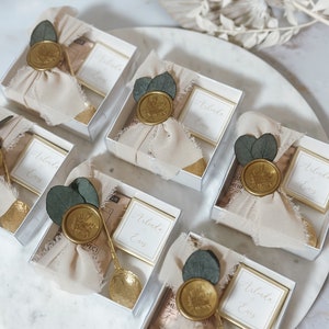 May include: Five small, clear gift boxes with white lids, each containing a small, white card with the text "Arlinda & Enes" printed in black. Each box is decorated with a gold wax seal, a small sprig of greenery, and a piece of beige fabric. The boxes are arranged in a row on a white surface.