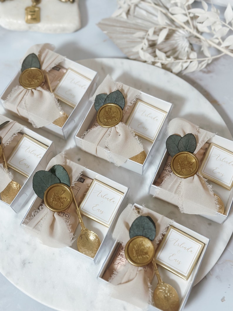 May include: Five small, clear gift boxes with white lids, each containing a small, white card with the text "Arlinda & Enes" printed in black. Each box is decorated with a gold wax seal, a small sprig of greenery, and a piece of beige fabric. The boxes are arranged in a row on a white surface.