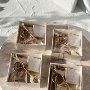 May include: Four small white boxes with clear lids, each containing a gold spoon, a small bag of brown tea, a small square of chocolate, and a gold wax seal with a leaf design. The boxes are tied with gold ribbon and each has a small white card with the text "Selune & Ali" written in black.