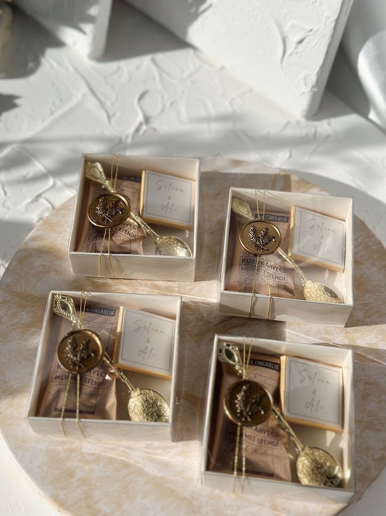 May include: Four small white boxes with clear lids, each containing a gold spoon, a small bag of brown tea, a small square of chocolate, and a gold wax seal with a leaf design. The boxes are tied with gold ribbon and each has a small white card with the text "Selune & Ali" written in black.