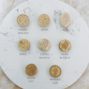 May include: Assortment of ten gold-toned wax seal stamps with various designs, including a laurel wreath, leaves, a rose, rings, a branch, an olive branch, a modern leaf, and the words "Thank You".