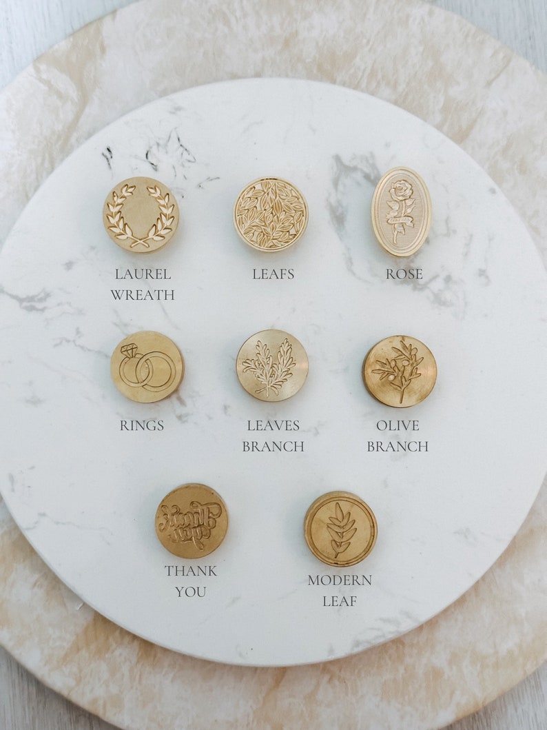 May include: Assortment of ten gold-toned wax seal stamps with various designs, including a laurel wreath, leaves, a rose, rings, a branch, an olive branch, a modern leaf, and the words "Thank You".