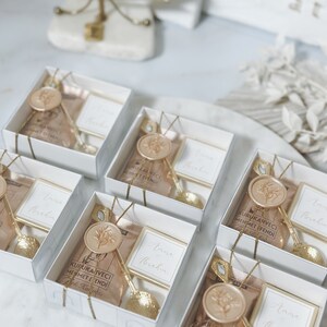 May include: A collection of small white gift boxes with gold accents. Each box contains a small bag of coffee, a gold spoon, and a card with the text "Anise Brakin" on it. The boxes are arranged in a circle on a white surface.