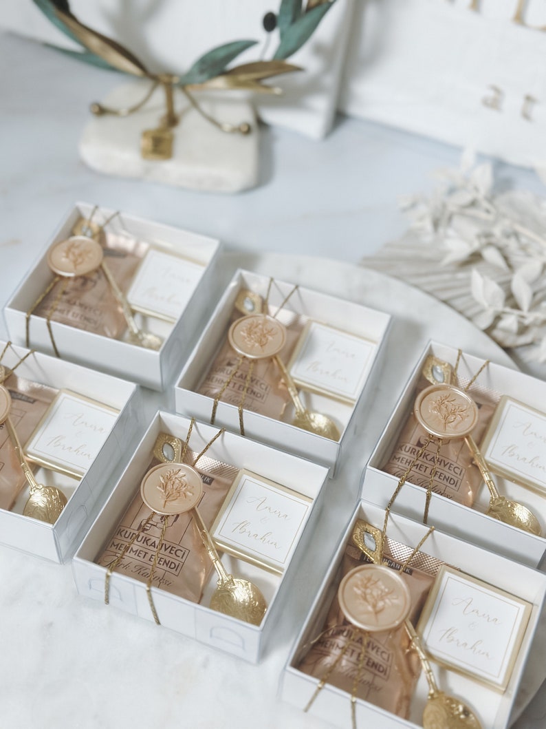 May include: A collection of small white gift boxes with gold accents. Each box contains a small bag of coffee, a gold spoon, and a card with the text "Anise Brakin" on it. The boxes are arranged in a circle on a white surface.