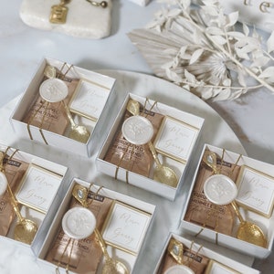 May include: Six small white boxes with clear lids, each containing a small gold spoon, a brown paper packet, and a white wax seal with a floral design. The boxes are tied with gold ribbon and have the text "Merve Gunay" on a white card inside each box.