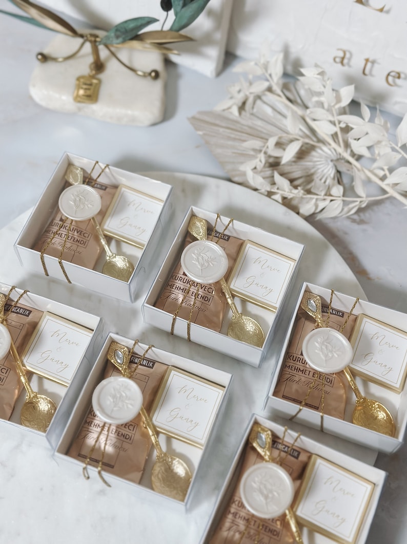 May include: Six small white boxes with clear lids, each containing a small gold spoon, a brown paper packet, and a white wax seal with a floral design. The boxes are tied with gold ribbon and have the text "Merve Gunay" on a white card inside each box.