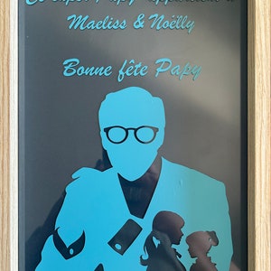 May include: A framed silhouette of a man with glasses and two children. The text "Ce super Papy appartient à Maeliss & Noëlly Bonne fête Papy" is written in blue on a dark gray background.