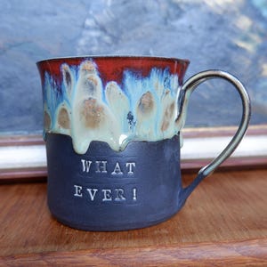 Könnte beinhalten: Eine Keramik-Tasse mit dunkelblauem Boden und rotem und blauem glasiertem Oberteil. Die Tasse hat die Worte "WHAT EVER!" in weißer Schrift. Der Griff ist silberfarben. Die Tasse steht auf einer Holzoberfläche.