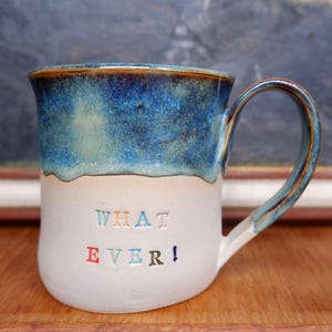 Könnte beinhalten: Eine weiße Keramik-Tasse mit schwarzem Henkel und dem Text "But First, Coffee" in fetter, schwarzer Schrift. Die Tasse steht vor einem verschwommenen Hintergrund, der eine gemütliche Umgebung suggeriert.