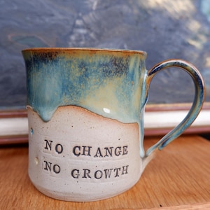 Könnte beinhalten: Ein Keramikbecher mit gesprenkelter cremeweißer Basis und einer glänzenden blau-grünen Tropfglasur im oberen Bereich. Der Text NO CHANGE NO GROWTH ist in schwarzen Buchstaben auf der Vorderseite eingeprägt. Der Becher hat einen geschwungenen Henkel.