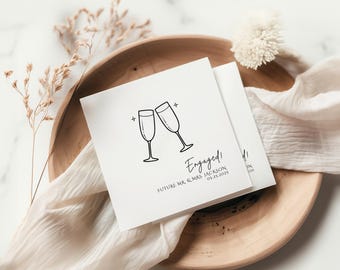 May include: White card with a black illustration of two champagne glasses clinking. The text "Engaged!" is written in black cursive with the date "05.23.2023" and the words "Future Mr. & Mrs. Jackson" below.