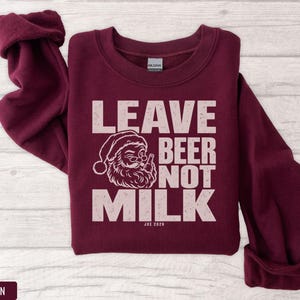 A maroon crewneck sweatshirt with pink text reading LEAVE BEER NOT MILK and a graphic of Santa drinking.