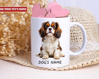 May include: White ceramic mug featuring a Cavalier King Charles Spaniel dog. The mug has the text "DOG'S NAME" at the bottom and "ADD YOUR PET'S NAME" at the top. Pink heart-shaped decorations are inside the mug.