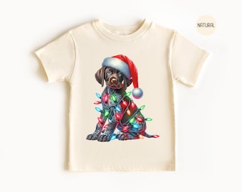 May include: A cream colored t-shirt with a graphic of a brown and black puppy wearing a red Santa hat and wrapped in Christmas lights.