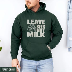 A man wears a forest green hooded sweatshirt with white text reading LEAVE BEER NOT MILK and a graphic of Santa Claus.